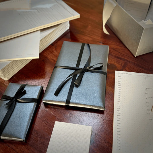 Gray gift boxes with black ribbons on a wooden surface with stationery items.