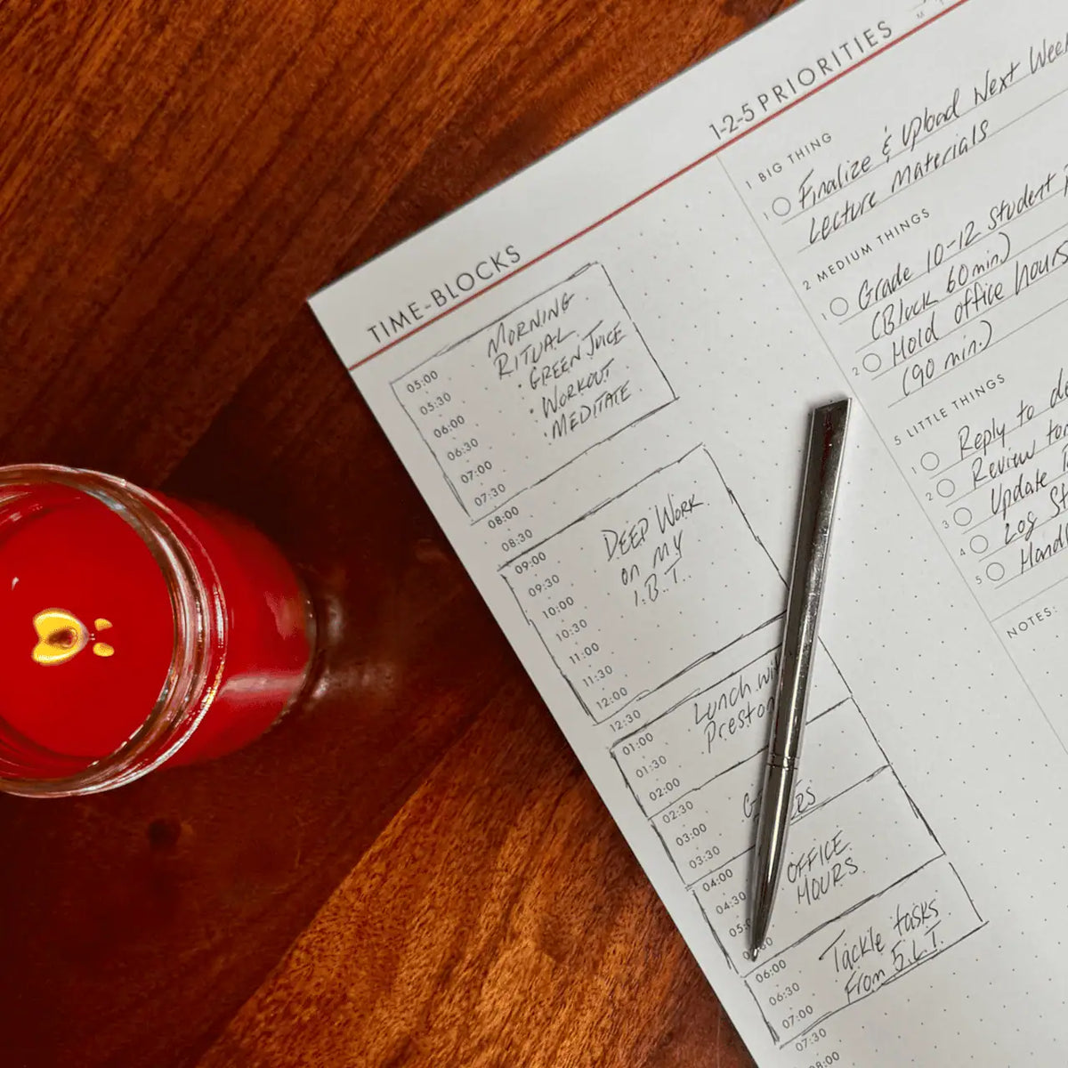 A close-up of a hand-written Time Block planner page titled '1-2-5 PRIORITIES' and 'TIME-BLOCKS' on a wooden desk, with a silver pen resting on the page and a red candle in a glass jar nearby.